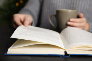 Woman with cup of beverage reading book at table, closeup